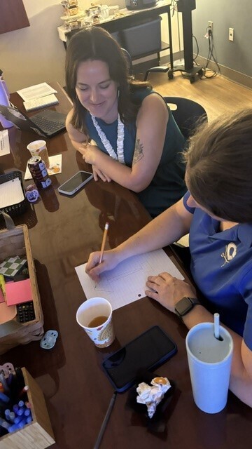 Two people sit at a desk writing on paper. One holds a pencil, the other a pen. Cups, a cell phone, a laptop, and a notepad are on the desk.
