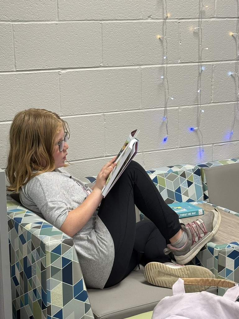A person in a gray shirt and glasses reads a book while sitting on a patterned couch.