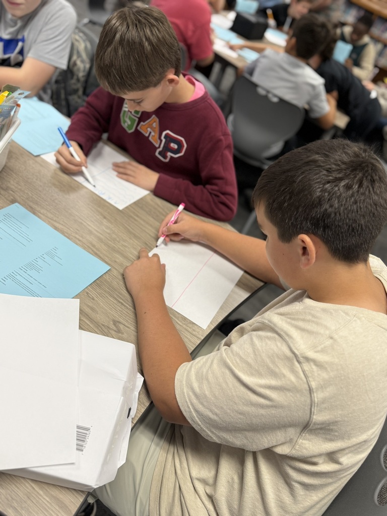 Two boys seated at a table, writing on paper with pens. One wears a maroon shirt, the other a white shirt.
