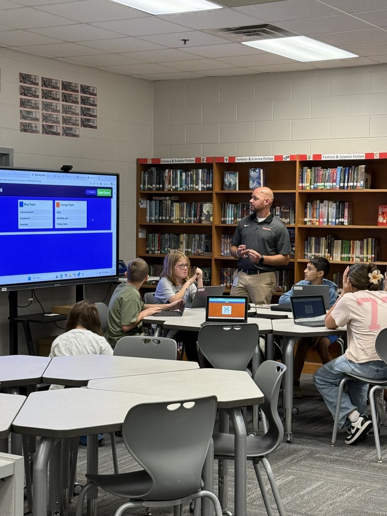 A teacher leads a group of students in a classroom. They sit at tables with laptops. Books are on shelves behind them.