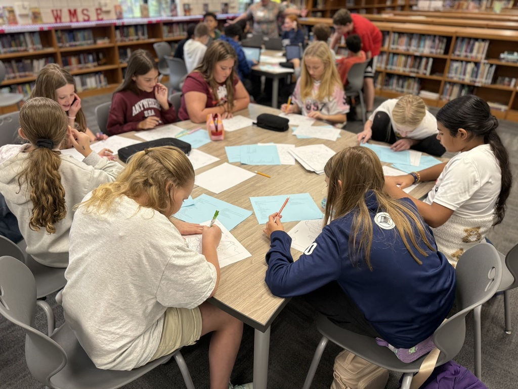 A group of students sit around a table in a library, writing on paper. Some wear jackets.
