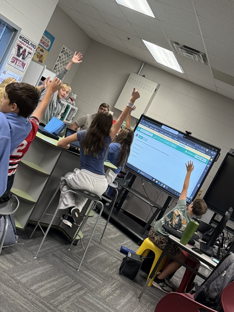 Classroom with children at tables. Some seated on stools, others standing, raising hands. Monitors and computer equipment on desks.