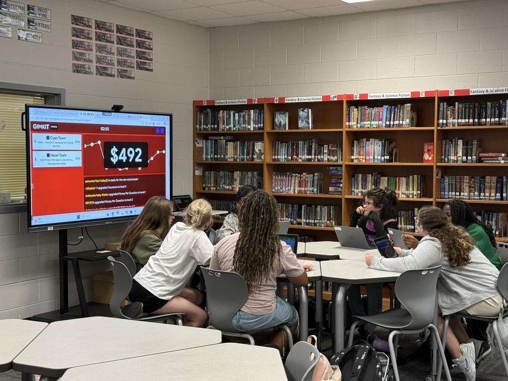 Students sit at desks in a library with a large monitor showing the text "$492". Shelves with books are in the background.