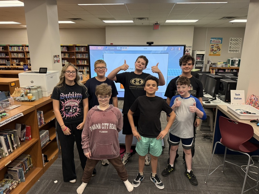 Group of children standing in a classroom with bookshelves and a large monitor behind them.