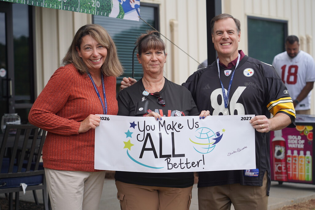 Three adults standing together holding a white sign that says, "You Make Us All Better!" against a blurred background.