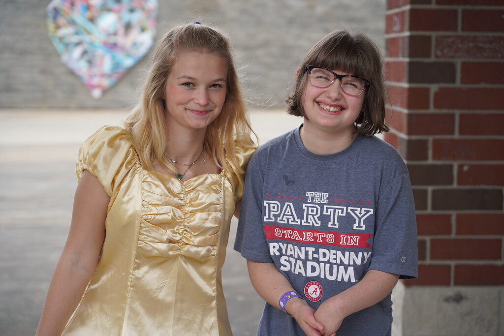 Two girls, one in a yellow dress and the other in a gray shirt, stand next to a brick wall.