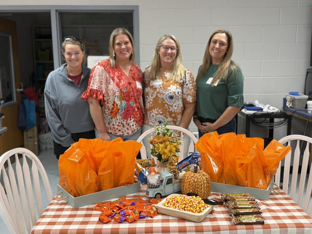 Four women stand behind a table with a checkered tablecloth. Orange bags and various candies are arranged.