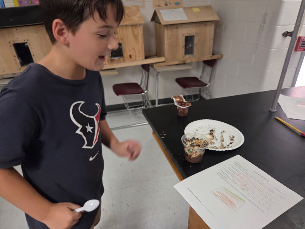 A boy stands in a classroom, holding a spoon and a paper, with a plate and cup behind him.