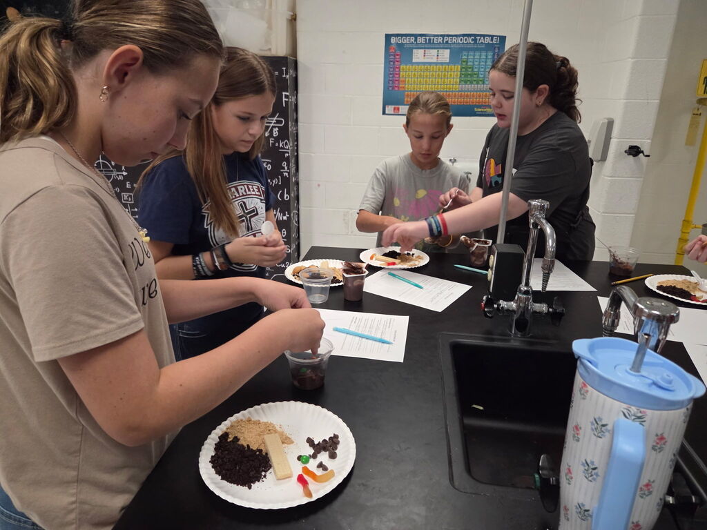 Four individuals, wearing casual clothes, stand at a lab table. Each person holds a paper plate with various food items and a cup.