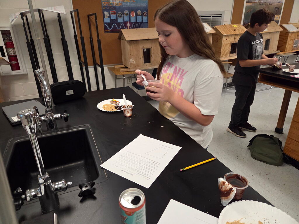 A student is standing at a counter, pouring liquid from a bottle into a cup.