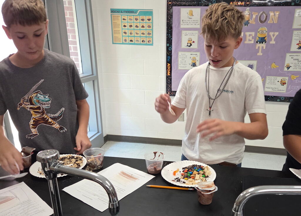 Two boys at a table, one holding a spoon and the other holding a cup. Papers, plates with food, and a sink are on the table.