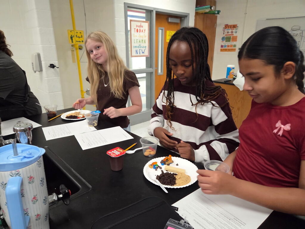 Three girls sit at a counter in a classroom, one girl eats from a plate, another holds a paper.