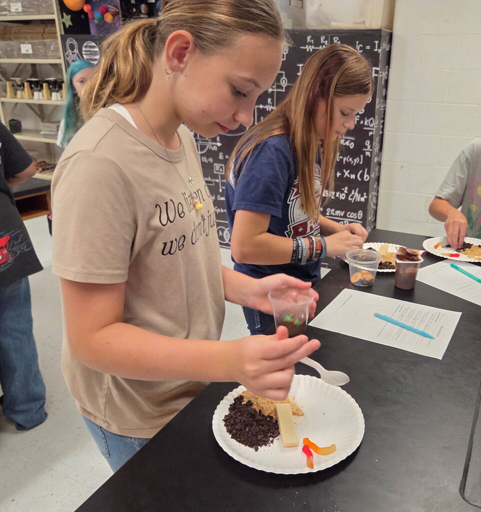 A girl adds ingredients to a plate with a cake. Another girl works behind her. A blackboard is in the background.