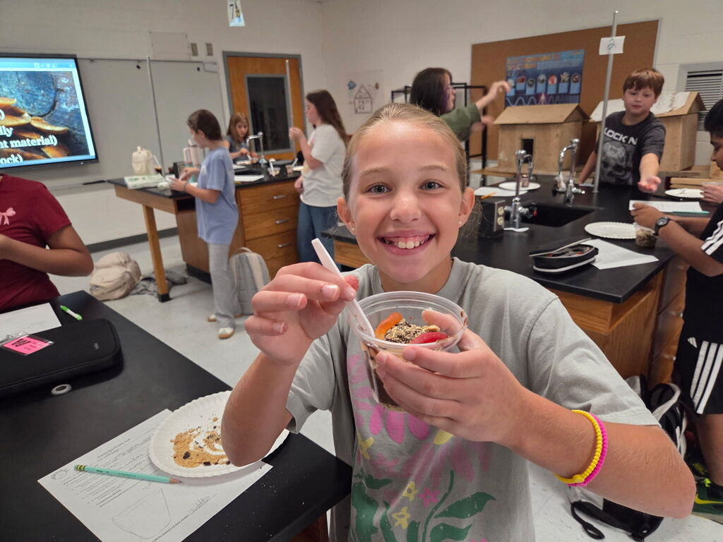A girl holds a bowl of food, smiling. Behind her, a classroom has tables and chairs. Students and adults work on tasks.