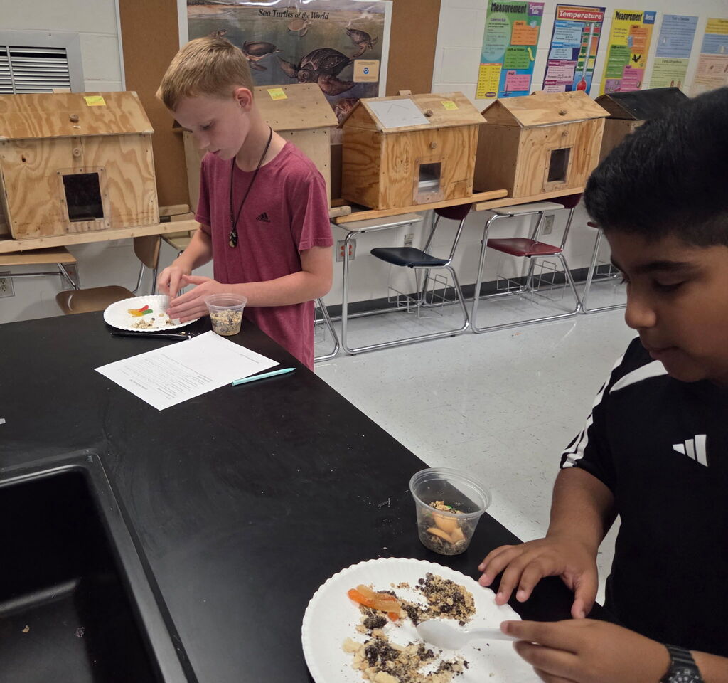 Two students in a classroom preparing food on plates. One student holds a spoon and another has a cup.