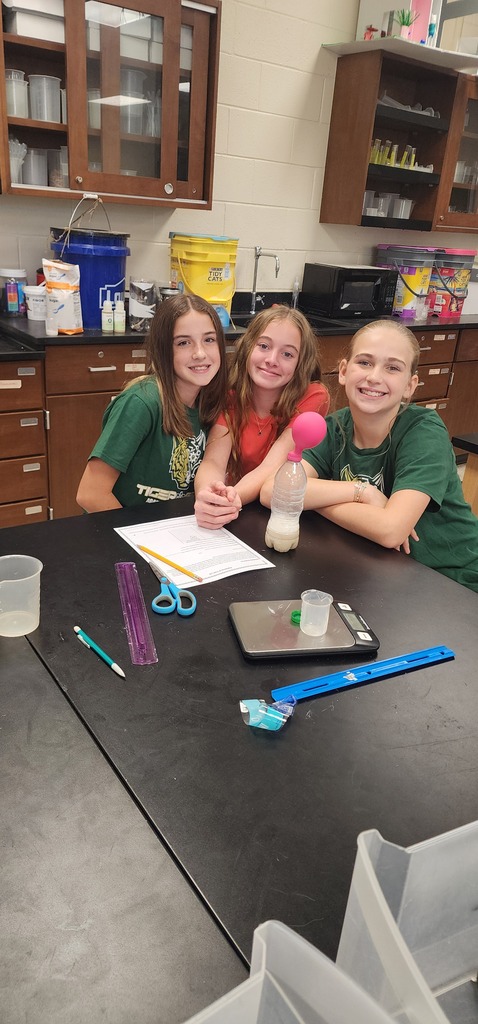 Three young people in green shirts work at a table with science equipment, in a room with wooden cabinets.