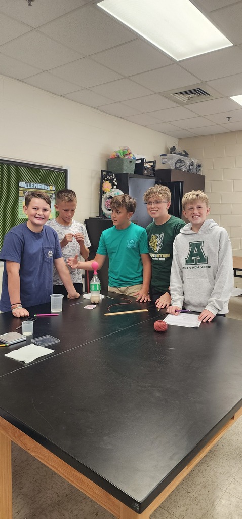 Five students are gathered around a long table, likely in a classroom. They are looking at a table with various items.