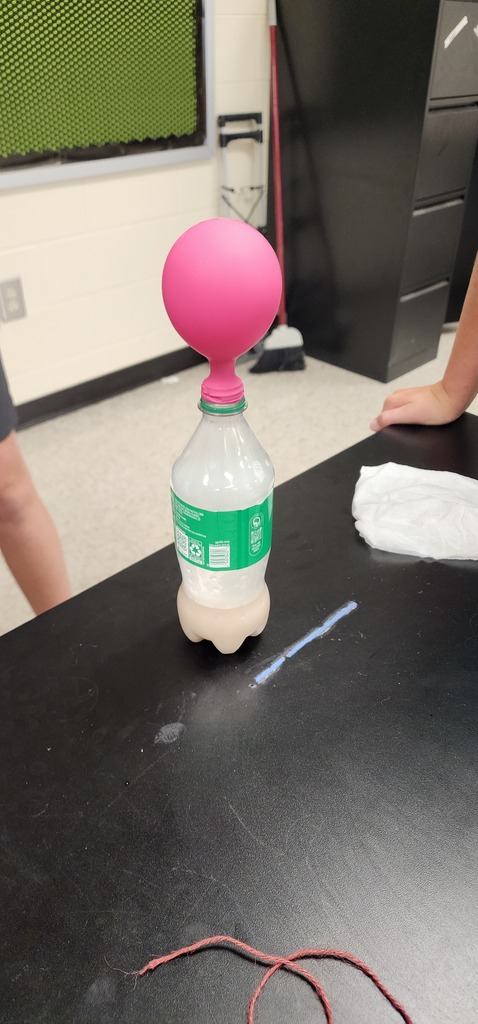 Pink balloon attached to a bottle cap and placed on a desk. Two people are present in the background.