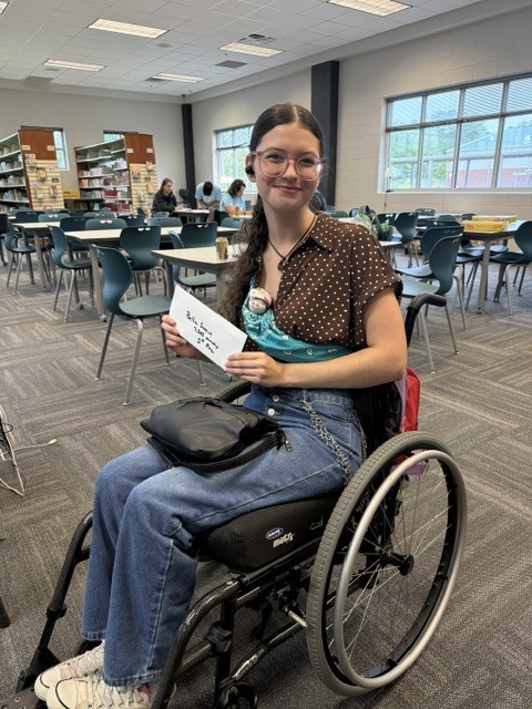 A woman in a wheelchair holds a paper in a room with tables, chairs, and bookshelves.