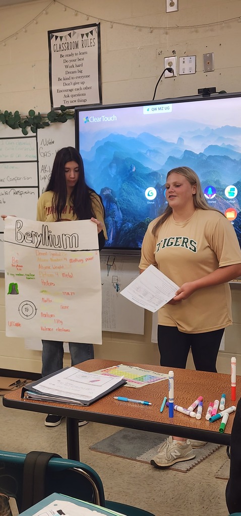 Two women hold posters in front of a large screen. A table holds papers, pens, and markers.