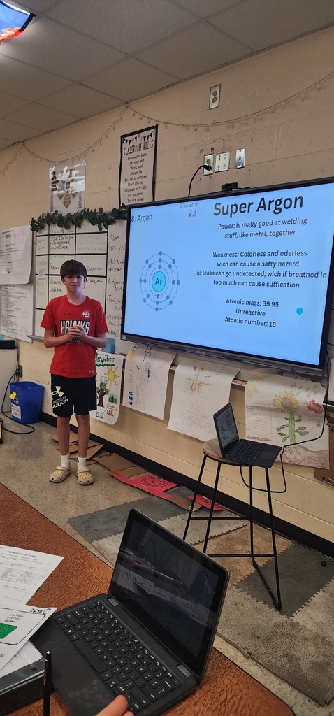 A person in a red shirt stands in front of a large screen projector with a presentation about argon.