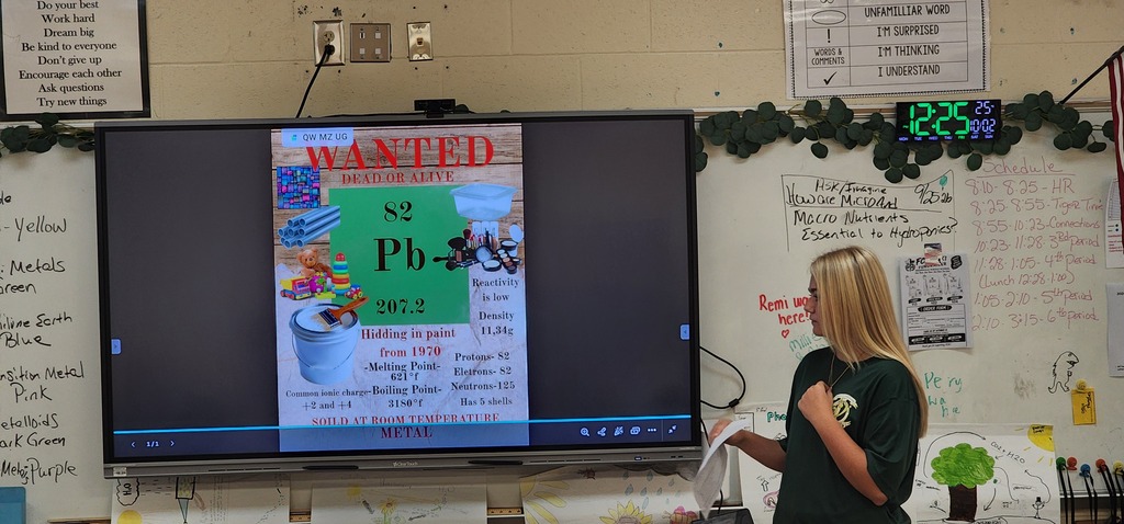 Woman stands by a classroom whiteboard with a monitor displaying a poster titled "Wanted: NP Ph-4".