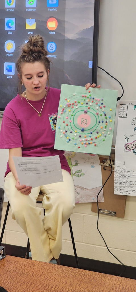 A student holds a paper and a colorful circular artwork in a classroom with a wall display and a screen behind.