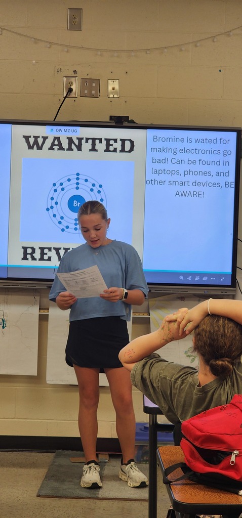 Person in blue shirt and black skirt reads aloud from paper in classroom. Student sits behind with head on desk.