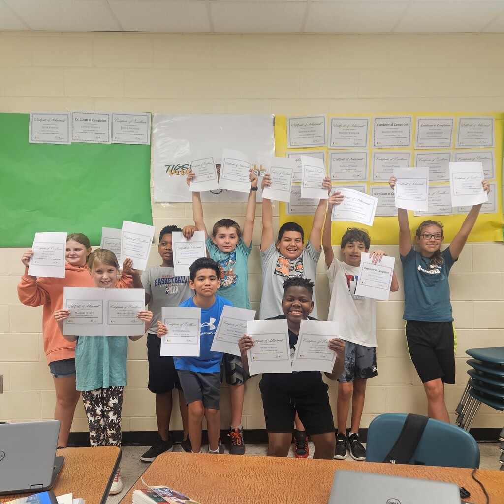 Group of children in a classroom holding certificates. They stand in a row, smiling. Papers are held high.
