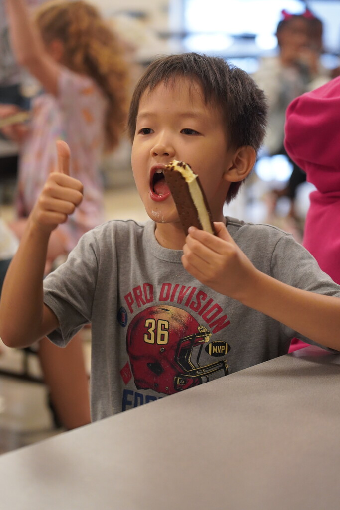 A young boy with a gray shirt eats an ice cream bar, giving a thumbs up. Other children are blurred in the background.