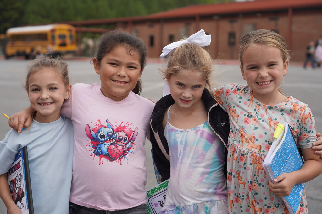 Four young girls, smiling, stand together in a school parking lot with a yellow school bus in the background.