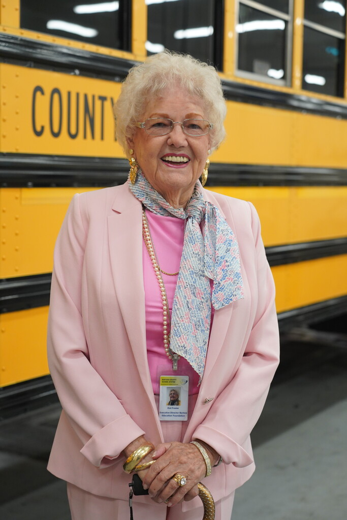 Woman with white hair wearing a pink suit and scarf stands in front of a yellow school bus.