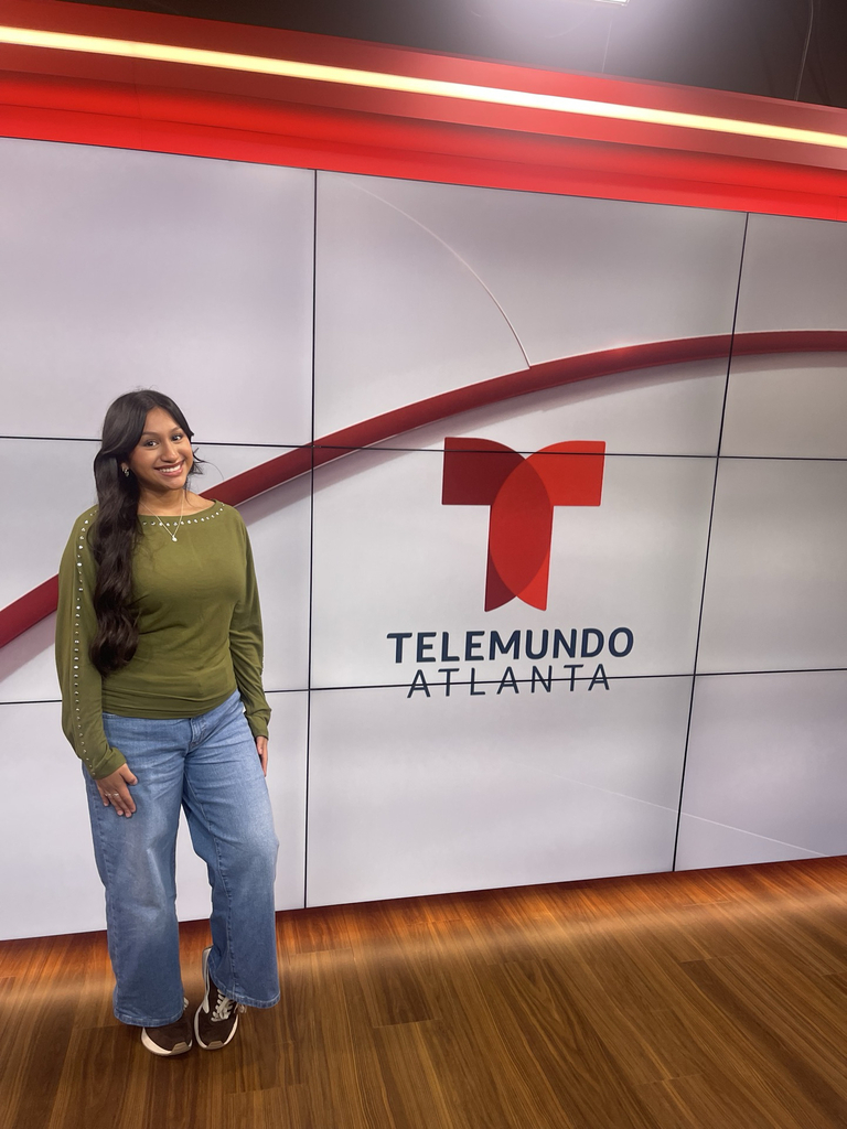 Person in green top and jeans stands in front of Telemundo Atlanta studio wall. Floor is wooden.