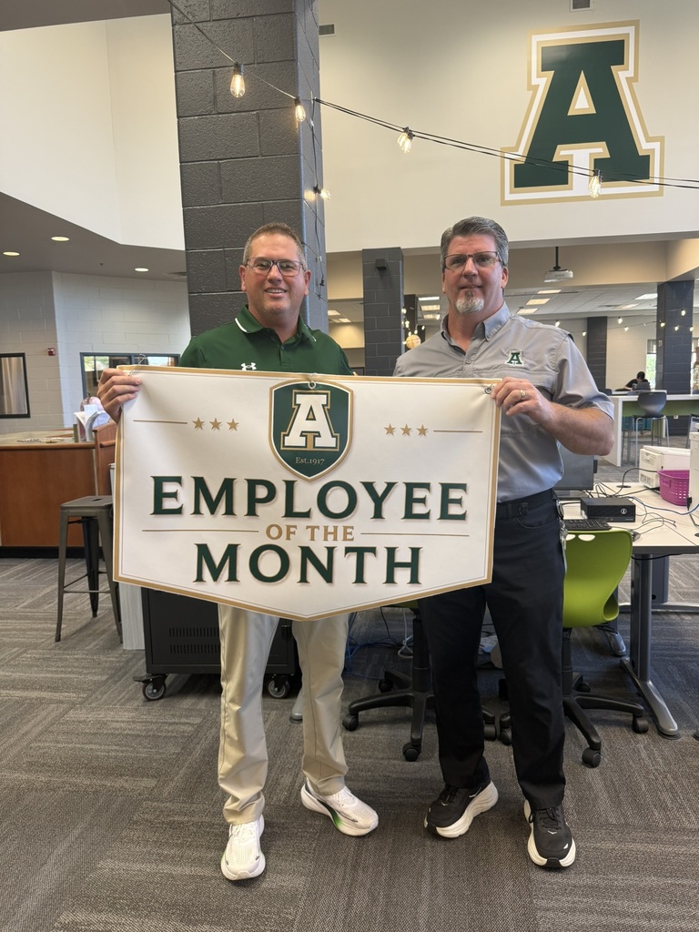 Two men stand in an office, holding a banner that reads "Employee of the Month". One wears a green polo, the other a gray polo.