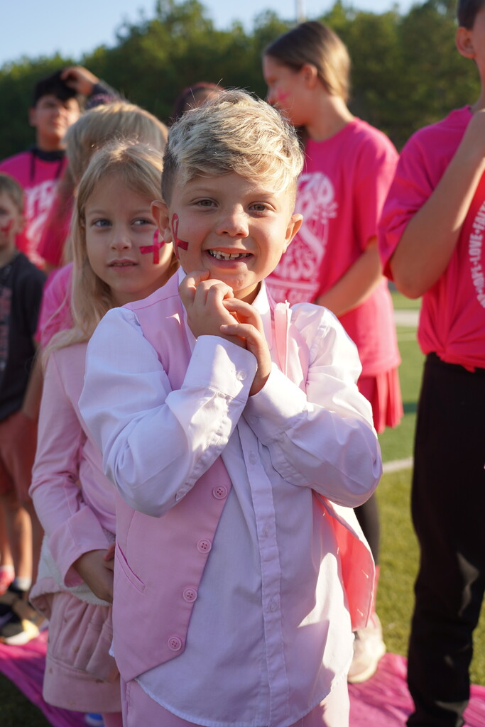 Children in pink shirts on a field, one with painted face, smiling, standing on pink carpet.