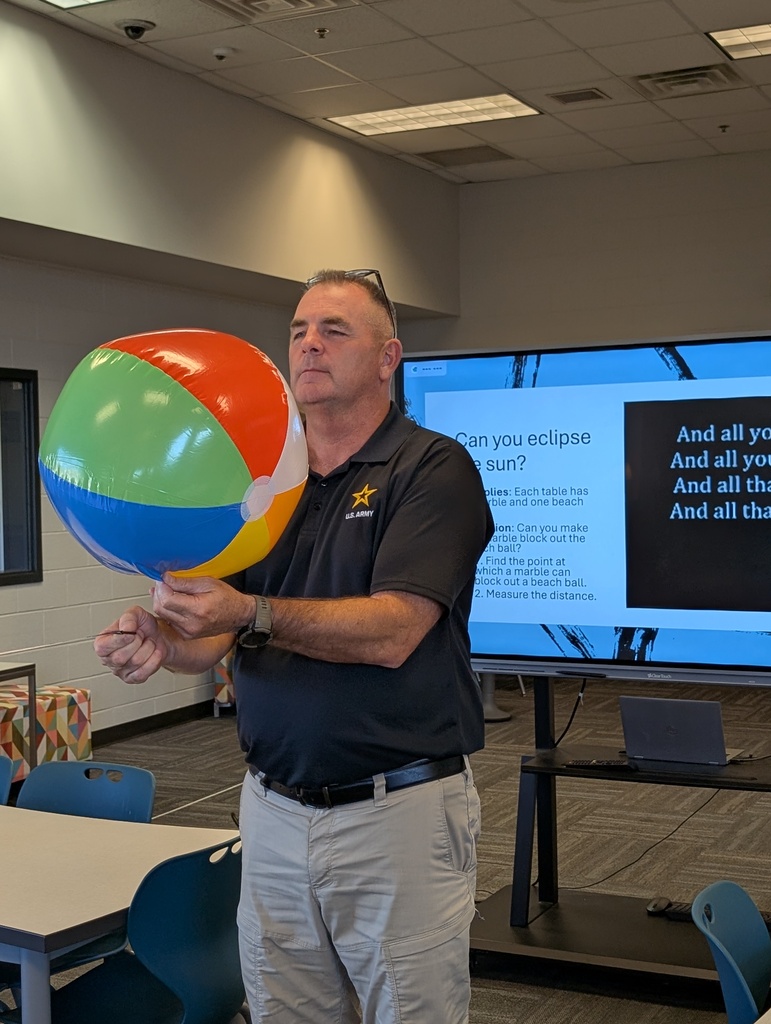 A teacher stands with a beach ball and tape measure as he attempts to determine at what distance the moon (a marble) blocks out the sun (a beach ball).