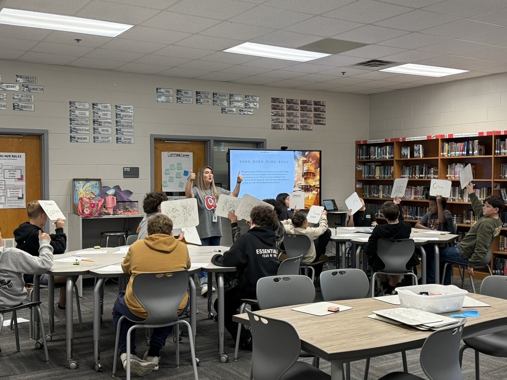 Classroom with rows of students at tables. A teacher stands at the front with a projector screen.