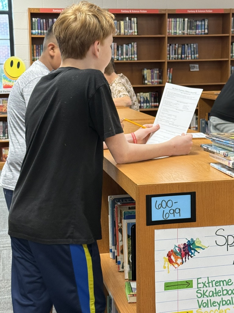 Several students in a library; one student reads from a paper in front of a desk.
