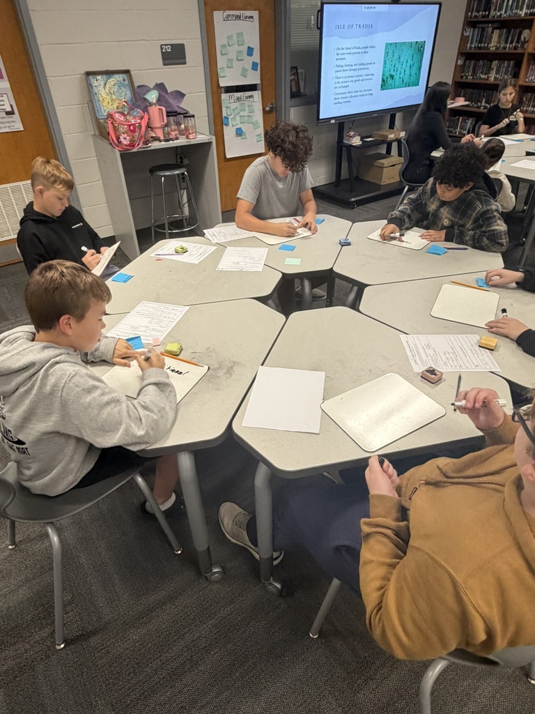 Students seated at round tables in a classroom, writing on papers with pens and pencils.
