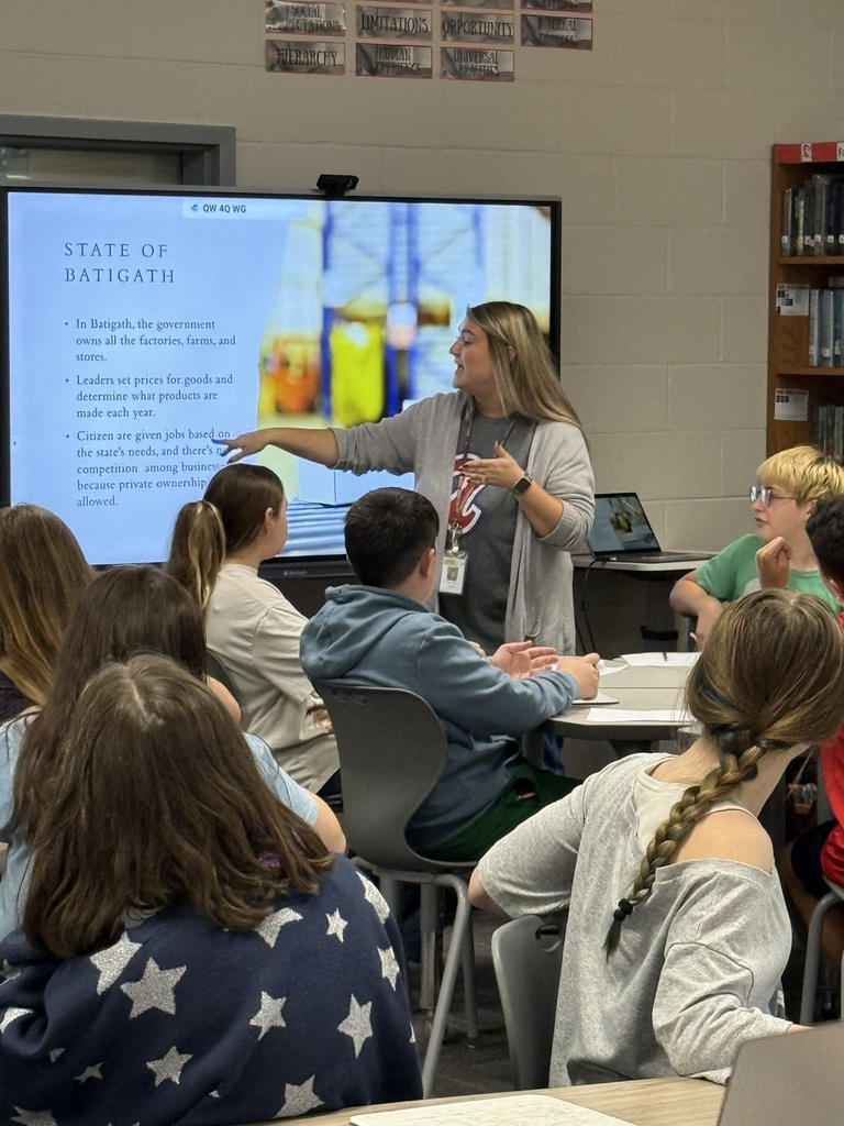 Woman with dark hair stands in front of screen displaying a presentation. Several students sit at tables.