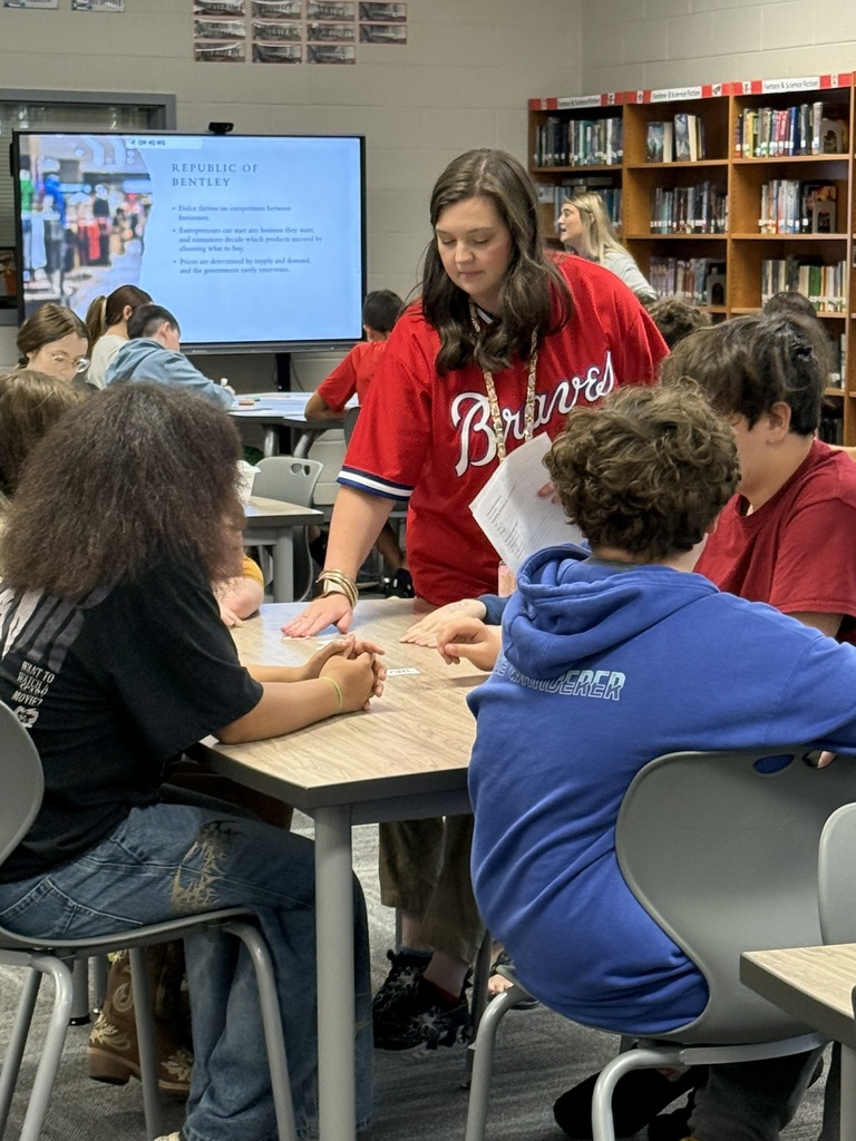 A woman stands next to a table where students sit in chairs. She holds a piece of paper.