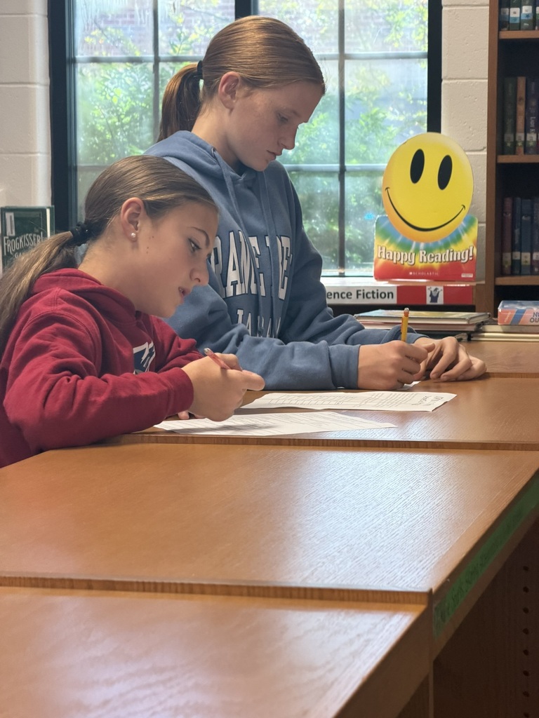 Two students in a classroom focus on writing with pencils at a wooden table, with a bookshelf in the background.