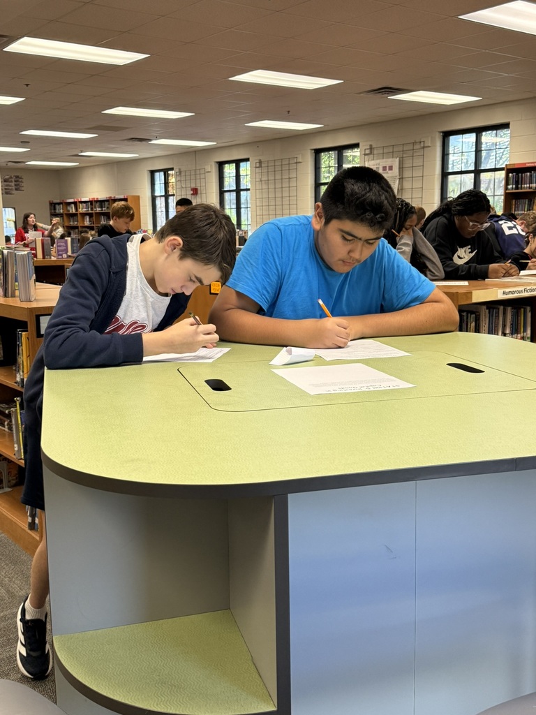 Two students seated at a table in a library, each writing on paper with pens.