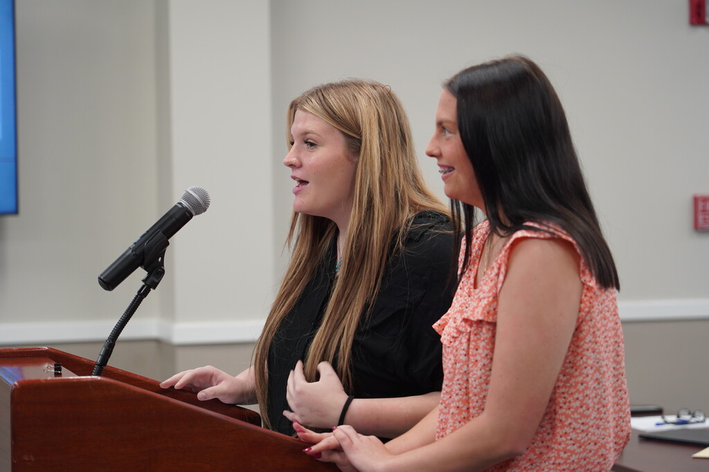 Two women speak into a microphone at a podium; one wears a black top, the other an orange blouse.