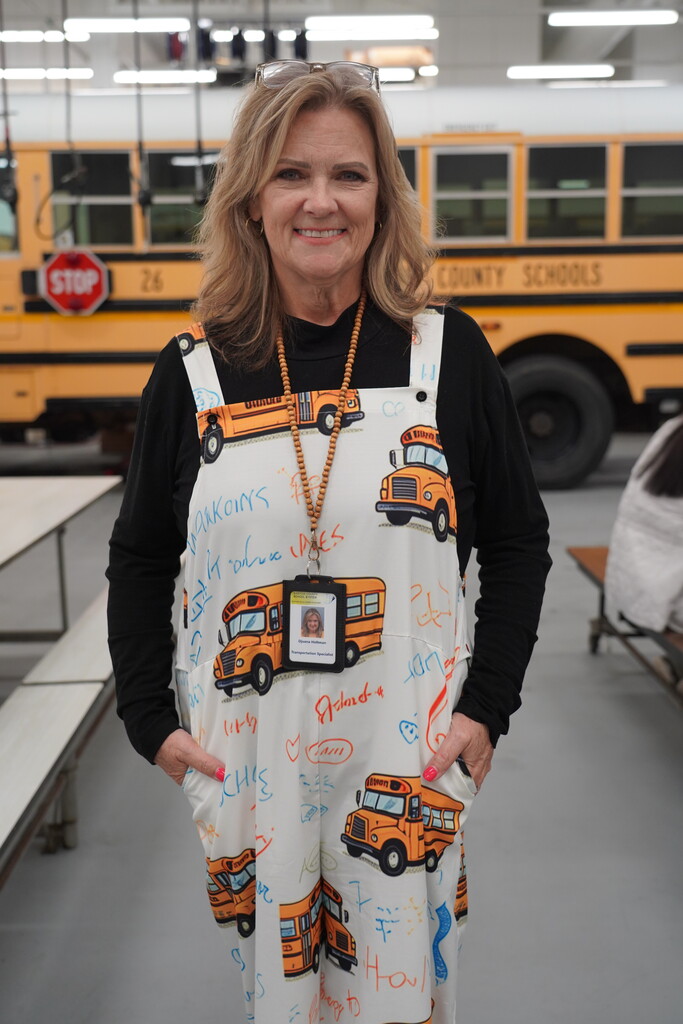 Woman in overalls with bus prints stands with hands on hips in front of a bus.