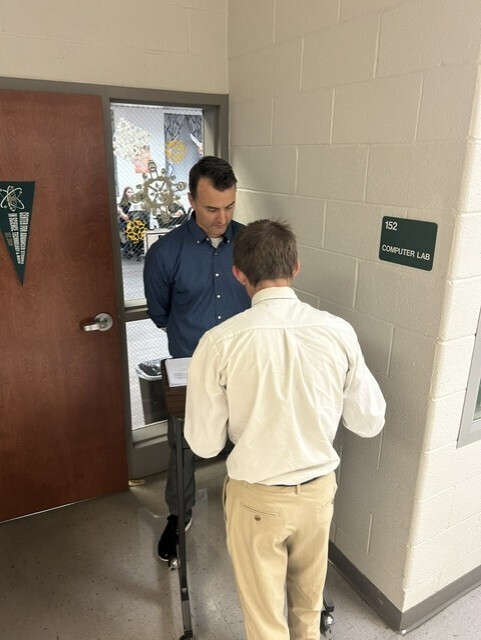 The picture shows a student and a teacher outside in the hall. The student is completing his formal 2-column proof and the teacher is assessing.