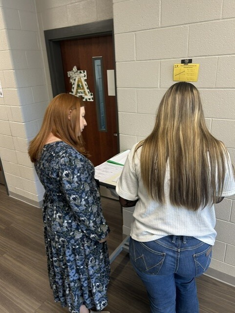The picture shows a student and a teacher outside in the hall. The student is completing his formal 2-column proof and the teacher is assessing.