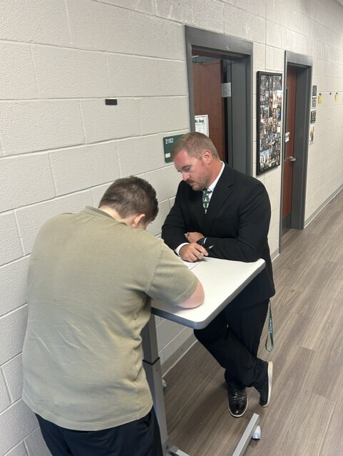 The picture shows a student and a teacher outside in the hall. The student is completing his formal 2-column proof and the teacher is assessing.