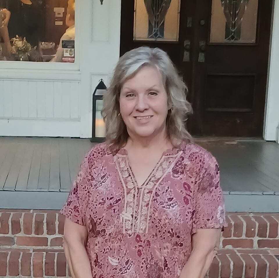 A woman with short gray hair stands in front of a brick wall. She wears a pink blouse and stands on a porch.