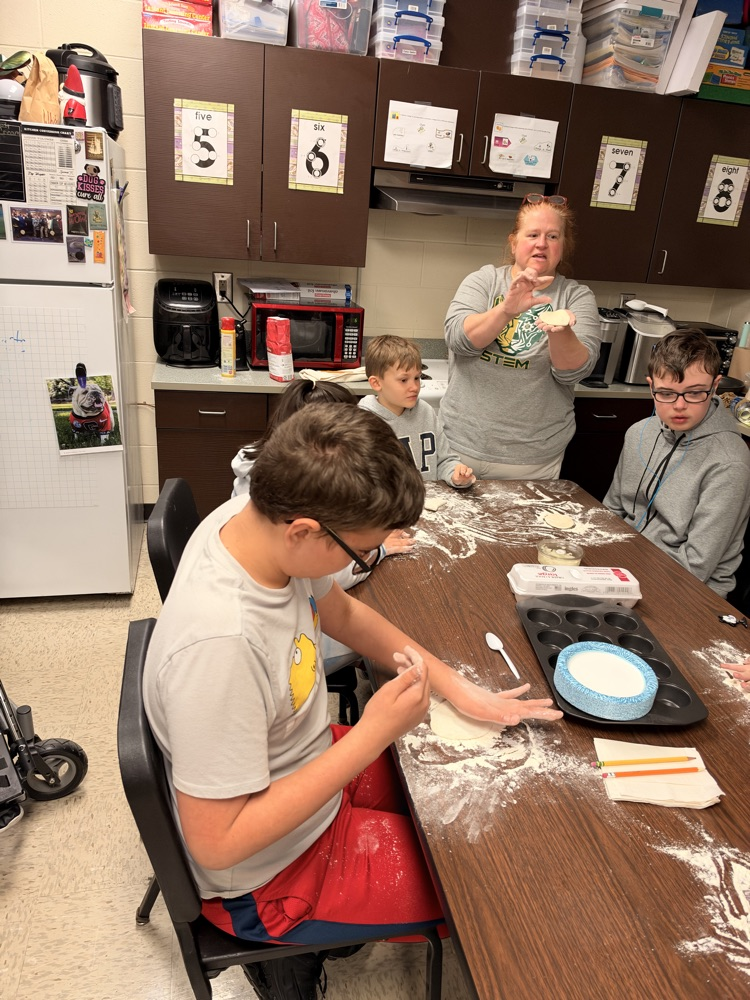 Kids seated around a table in a kitchen, using spoons to mix dough, with flour scattered around.